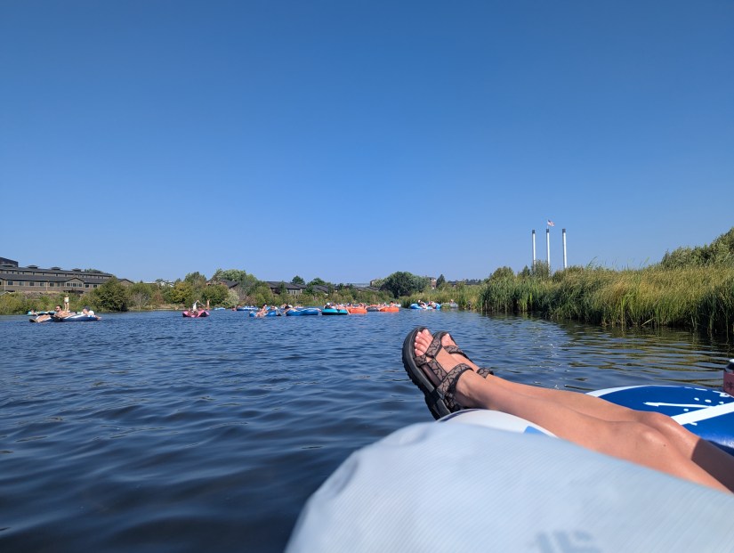 Scenic view of a river with multiple people floating on inner tubes, a pair of feet resting on the edge of a tube in the foreground, surrounded by greenery and a clear blue sky.