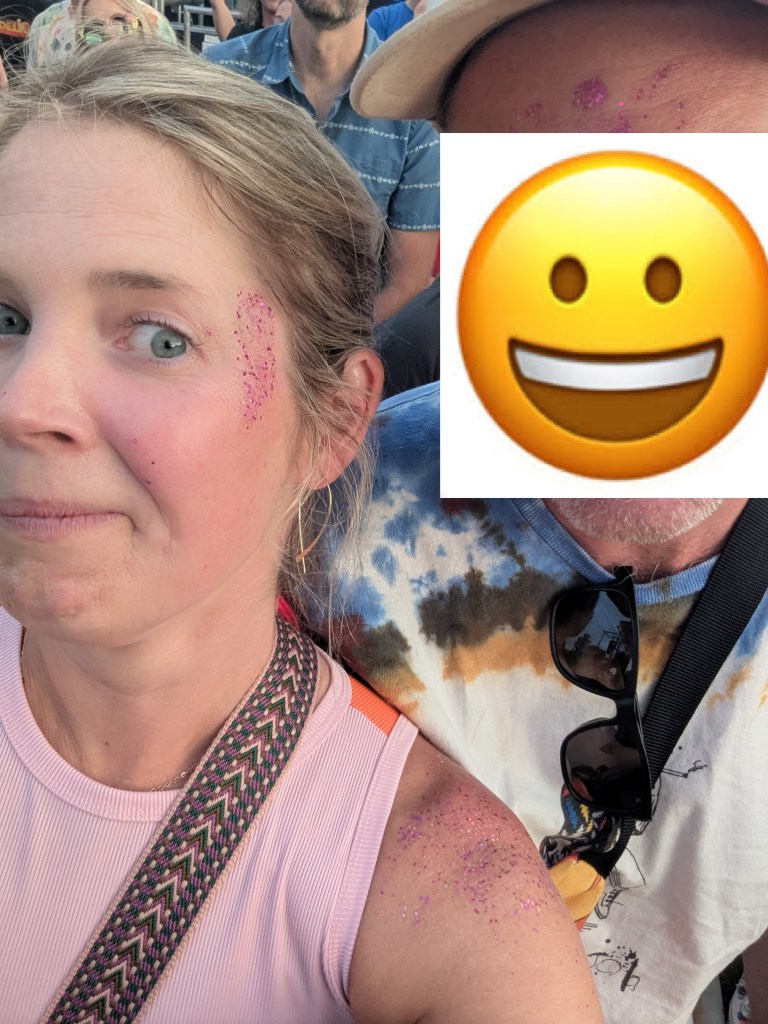 Close-up selfie of a woman with glitter on her face and shoulder, posing with a blurred figure beside her at a crowded event.