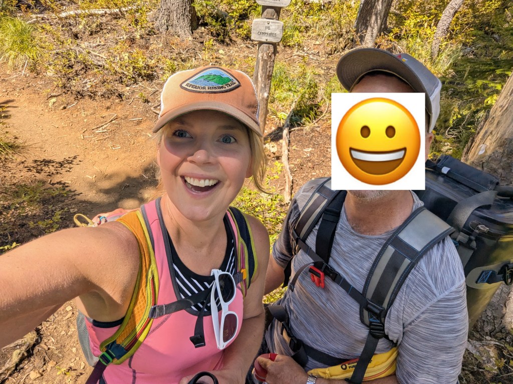 A smiling woman takes a selfie with a man while hiking on a trail. Both are wearing outdoor gear and appear happy in a natural setting with trees in the background.