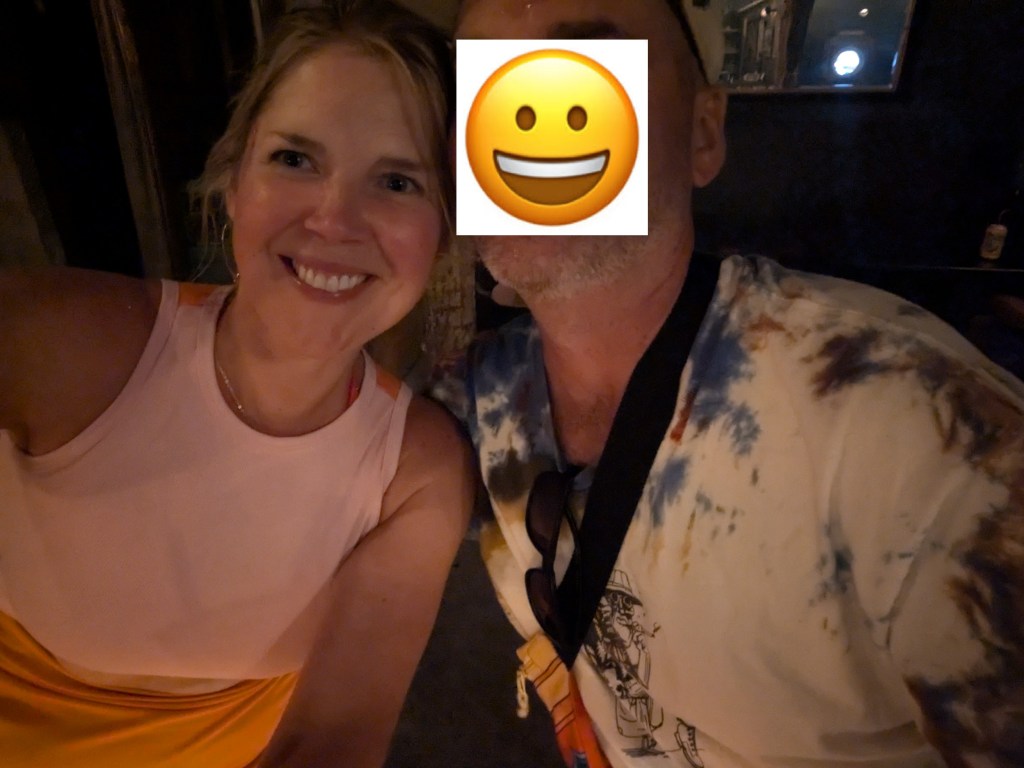 A woman smiling while taking a selfie with a man who has his face obscured. The woman is wearing a pink top, and the man is wearing a colorful tie-dye shirt.