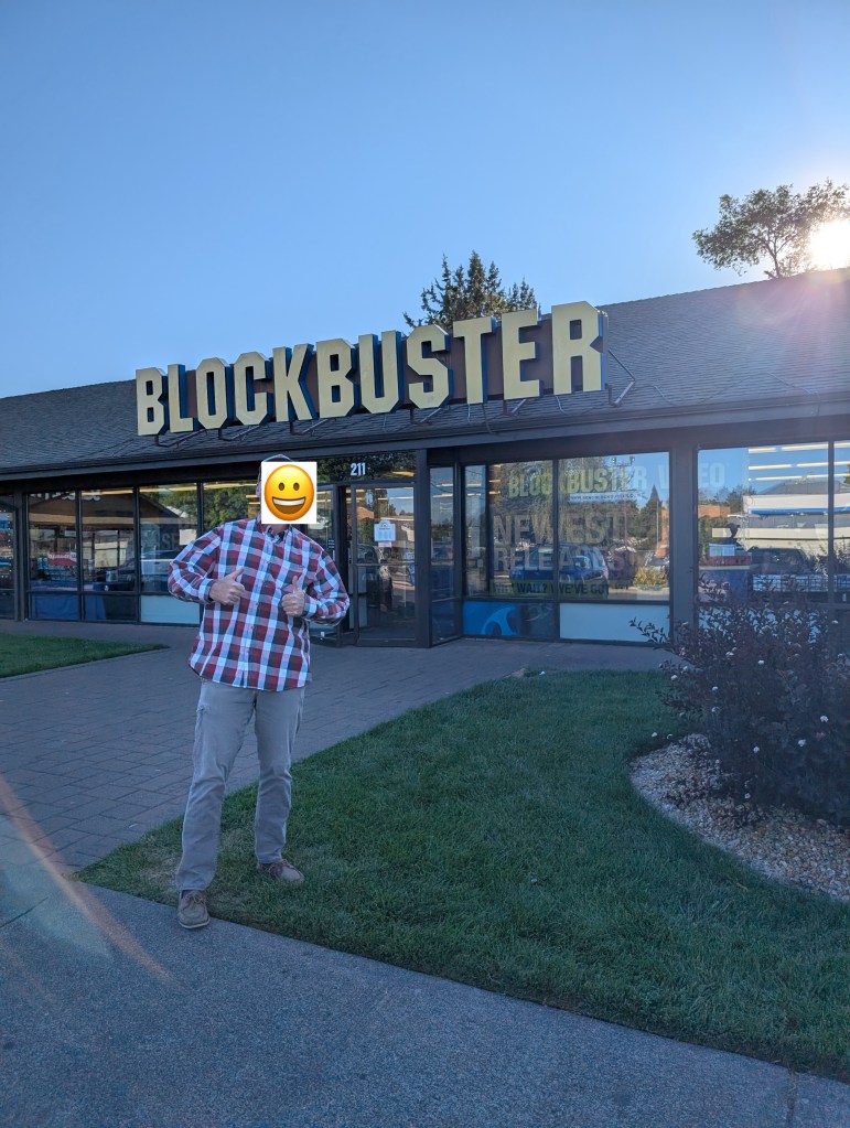A person in a plaid shirt stands outside a Blockbuster store, giving a thumbs up. The store sign is prominently displayed above the entrance.
