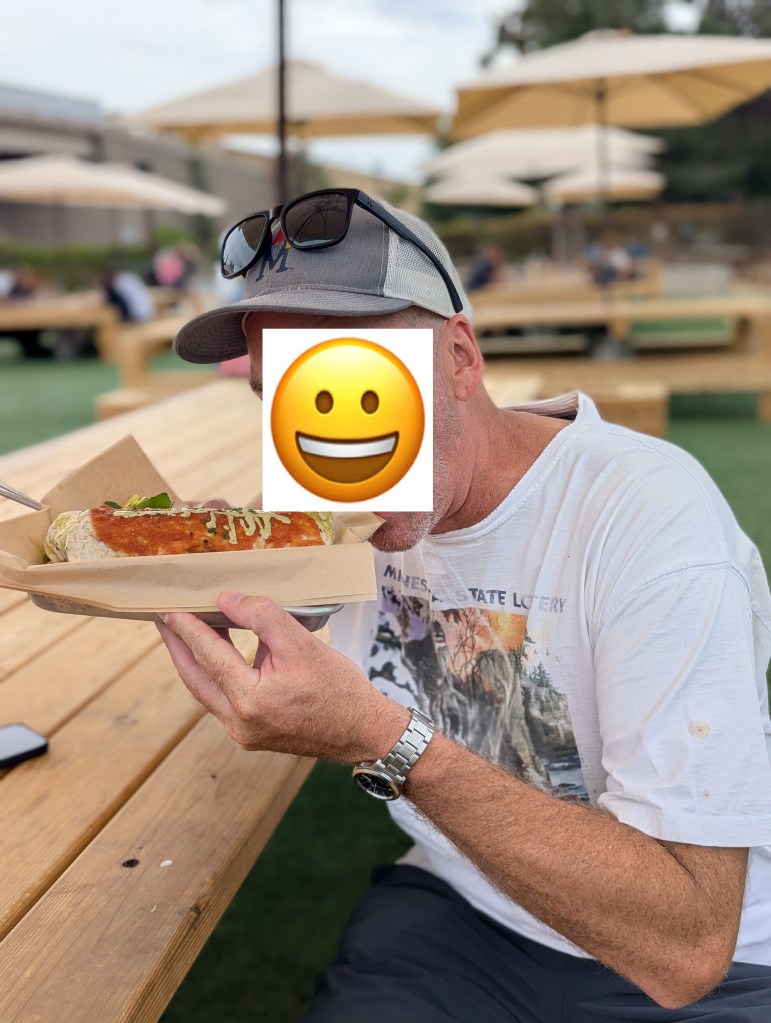 A person sitting at a picnic table outdoors, holding a large wrapped food item with toppings, wearing sunglasses and a casual t-shirt, in a lively outdoor setting with umbrellas.