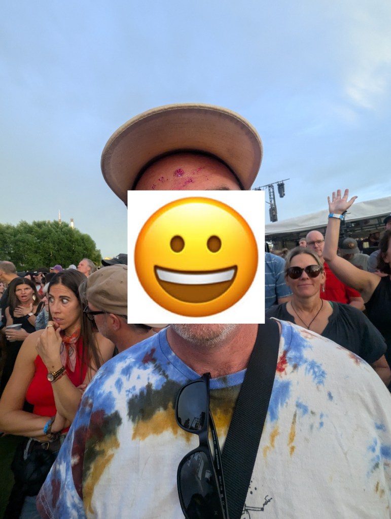 A group of people at a concert with a man in the foreground wearing a tie-dye shirt and sunglasses. The background features attendees enjoying the event.
