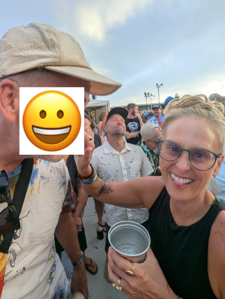A woman smiling and holding a metal cup in a crowded outdoor event, with several people in the background. The scene is lively and upbeat.