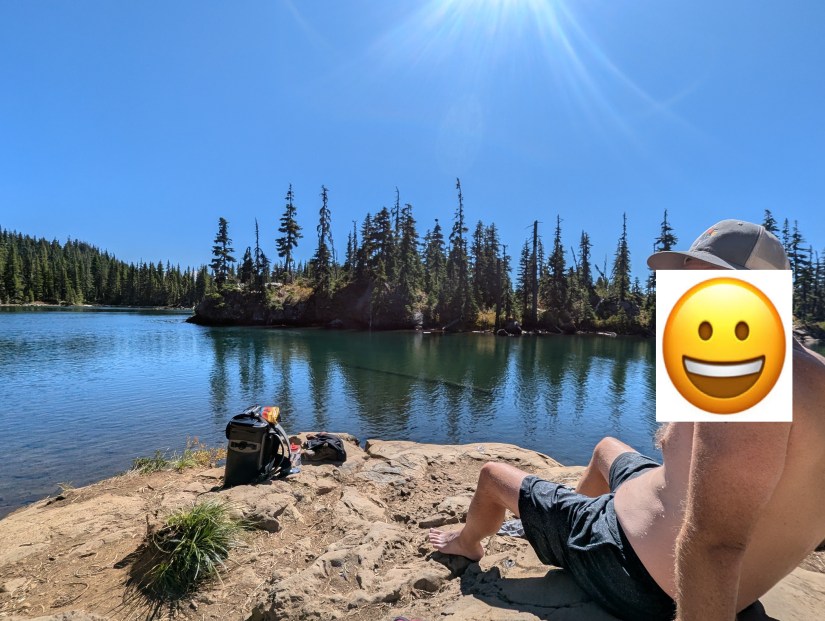 A person sitting on a rock by a tranquil lake surrounded by trees under a clear blue sky.