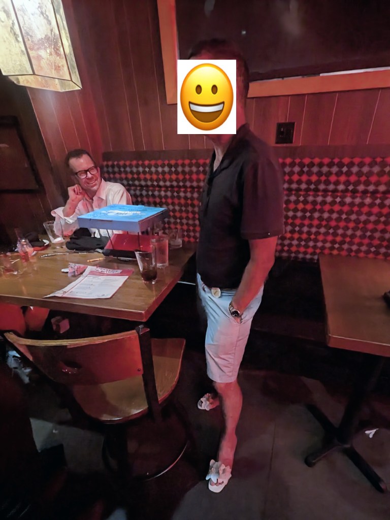 A man standing in a restaurant wearing a black shirt and light shorts, with flip-flops, while another man in the background sits at a table with drinks.