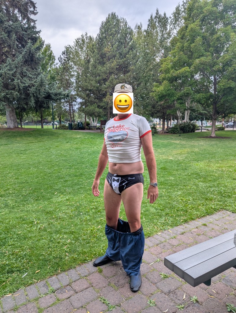 A person standing in a park wearing a Lake Superior t-shirt and black tuxedo-themed underwear, with pants pulled down to their ankles, and a smiling emoji covering their face.