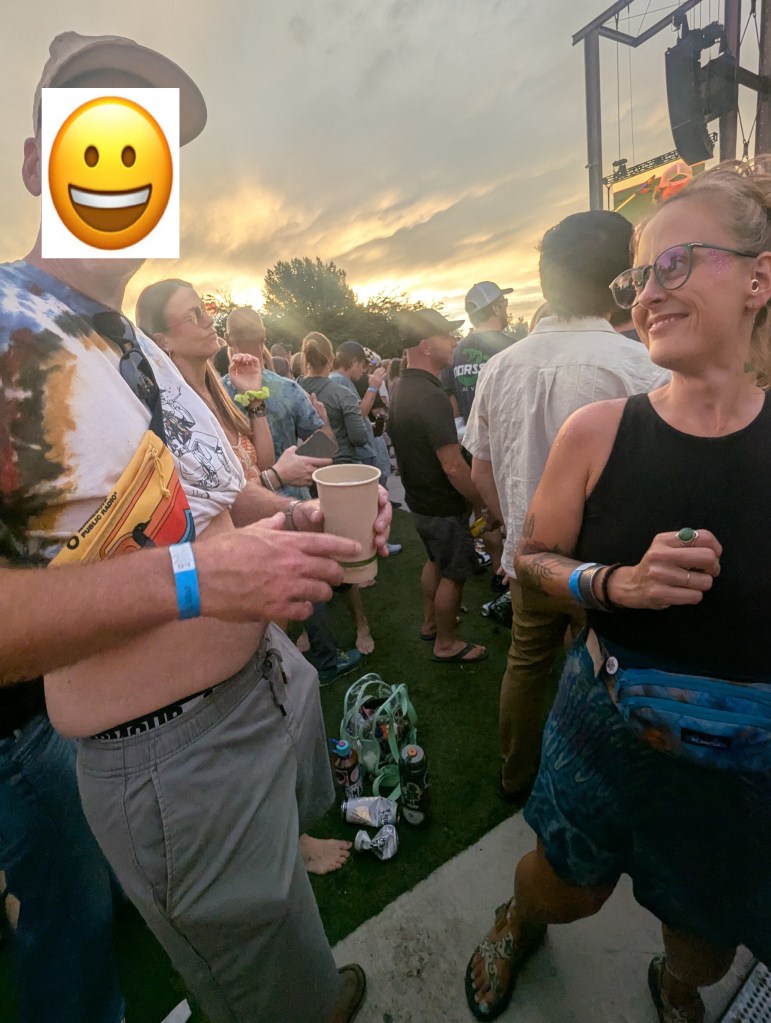 A group of people enjoying an outdoor concert at sunset, with one person wearing a colorful tie-dye shirt and holding a drink while another woman with glasses smiles nearby.