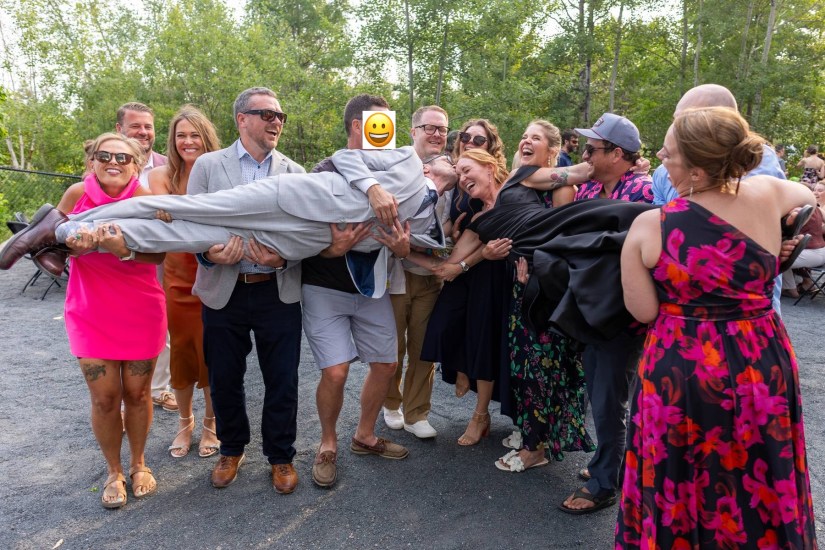 A lively group of people celebrating outdoors, with one person being playfully lifted and held by others. The scene is filled with joy and laughter, surrounded by greenery.