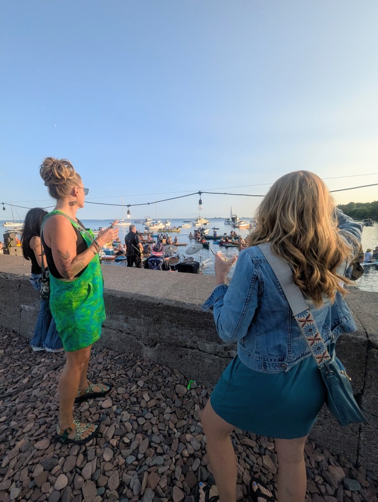 Two women standing by a waterfront, enjoying drinks and watching boats on the water during sunset.