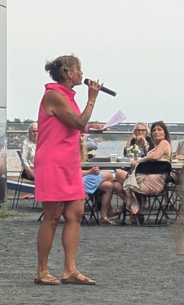 A woman in a bright pink dress speaks into a microphone at an outdoor event, while seated guests listen intently.