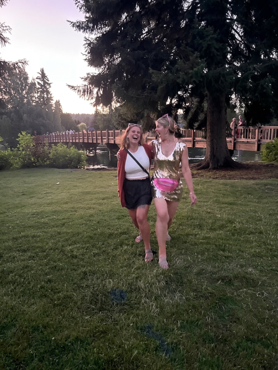 Two joyful women walking together on grass, smiling and laughing, with a wooden bridge and trees in the background during sunset.