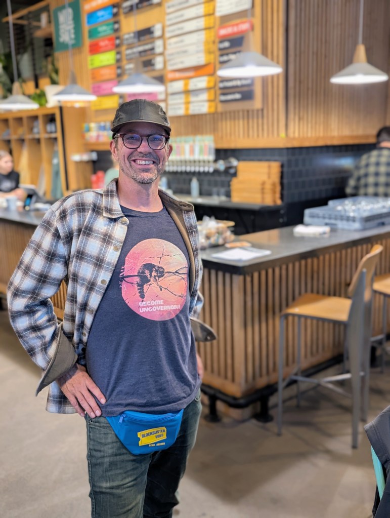 Man smiling and posing in a casual setting, wearing a plaid jacket, a graphic t-shirt, and a blue fanny pack, with a modern cafe background featuring a wooden bar and menu board.