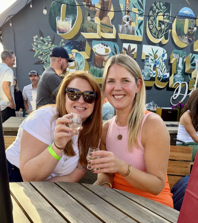 Two women smiling and holding small glasses, enjoying a drink at an outdoor festival. A vibrant mural with the text 'Oregon' is visible in the background, along with people socializing.