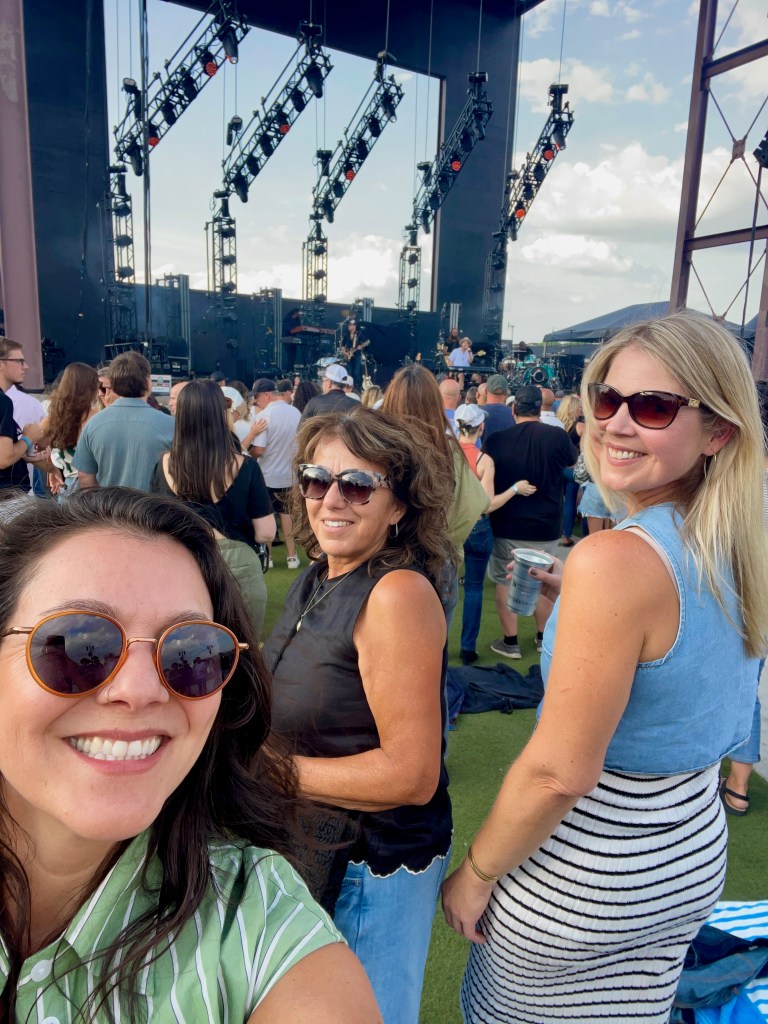 A group of three women taking a selfie at an outdoor concert, with a crowd in the background and a stage featuring lighting equipment.