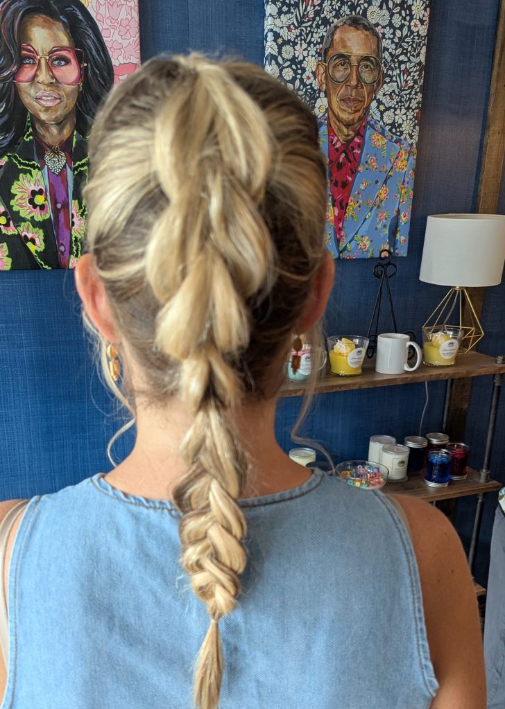 A woman with blonde hair styled in a braided ponytail, viewed from behind, standing in front of colorful portraits and a shelf with decorative items.