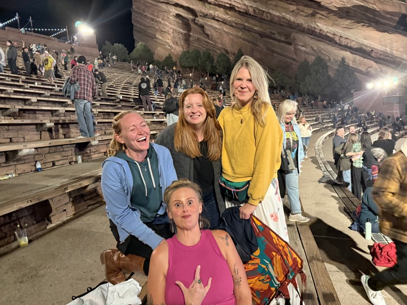 Four friends posing for a photo at a concert venue, smiling and enjoying their time together. The setting features an outdoor amphitheater with a rocky backdrop and spectators in the background.