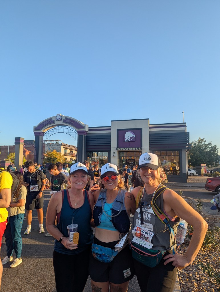 Three women posing together at an outdoor event in front of a Taco Bell restaurant, wearing hats and athletic attire.