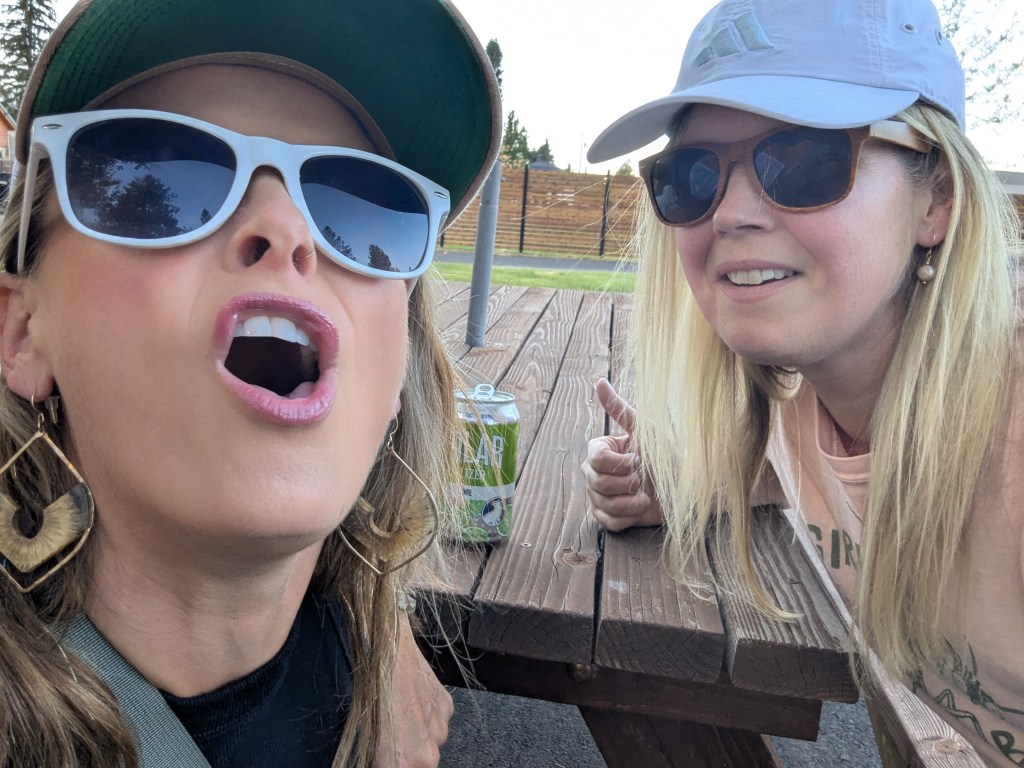 Two women posing playfully, one with sunglasses making a surprised facial expression and the other smiling and giving a thumbs up, sitting at a wooden picnic table outdoors.