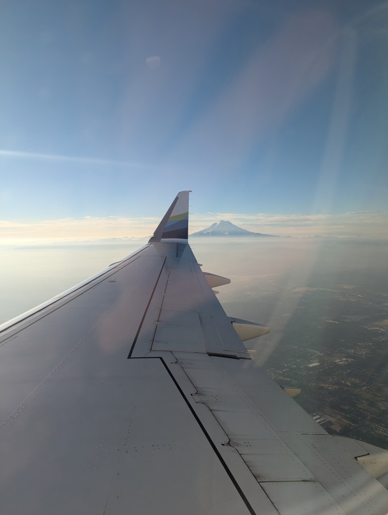 View from an airplane window featuring the aircraft wing and a distant mountain in the background under a clear blue sky.