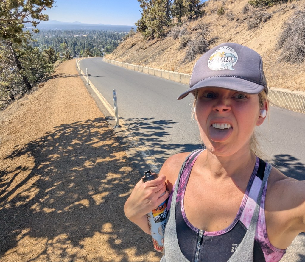 A woman taking a selfie on a winding road, making a funny face and holding a water bottle. Trees and hills are visible in the background under a clear blue sky.