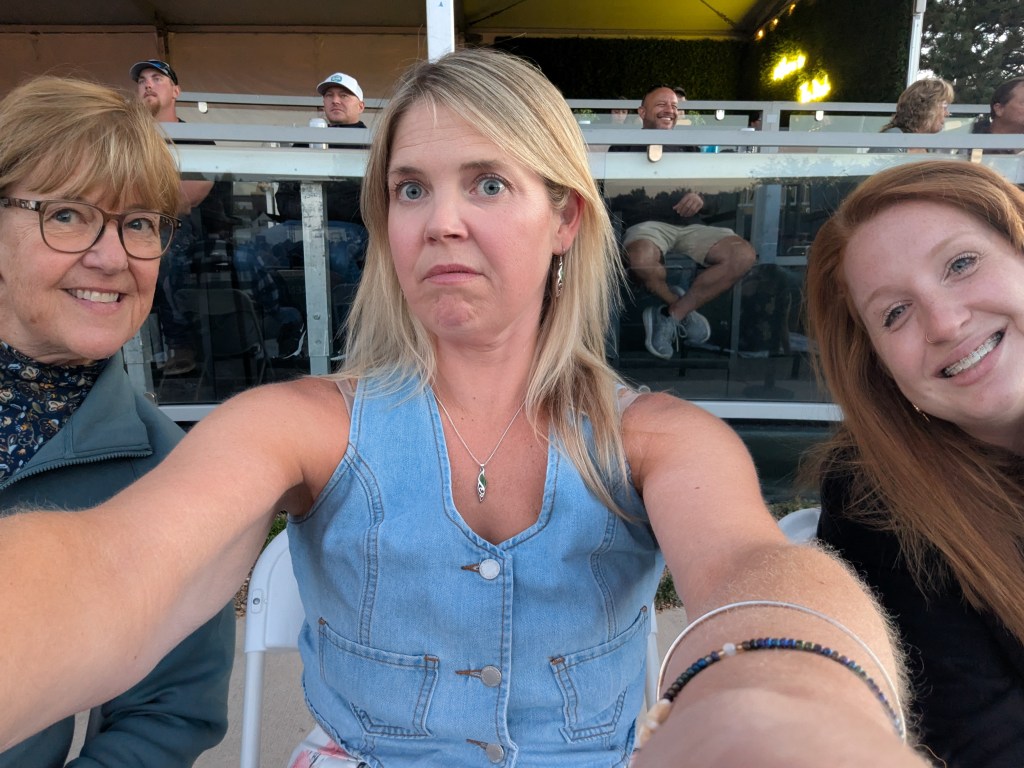 A group selfie of three women sitting together, two smiling and one making a playful expression. In the background, spectators can be seen seated behind them.