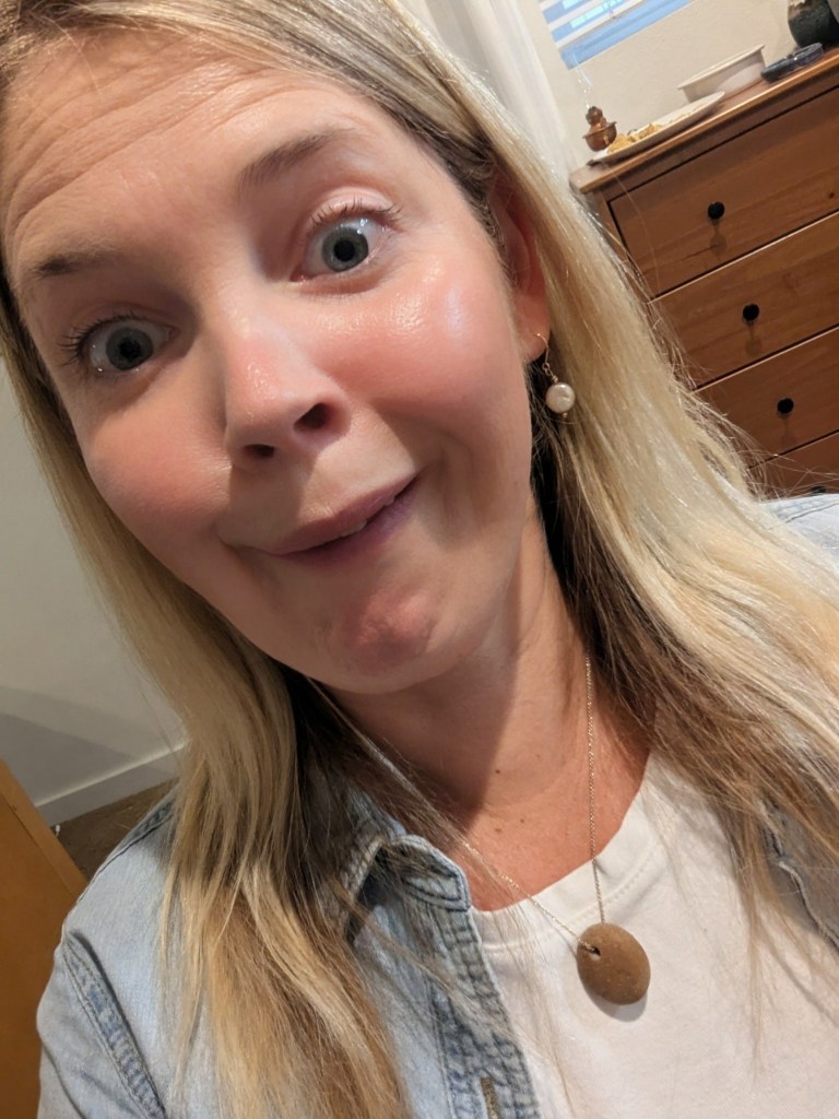 A woman making a humorous facial expression with wide eyes and a quirky smile, wearing a denim jacket and a white shirt.