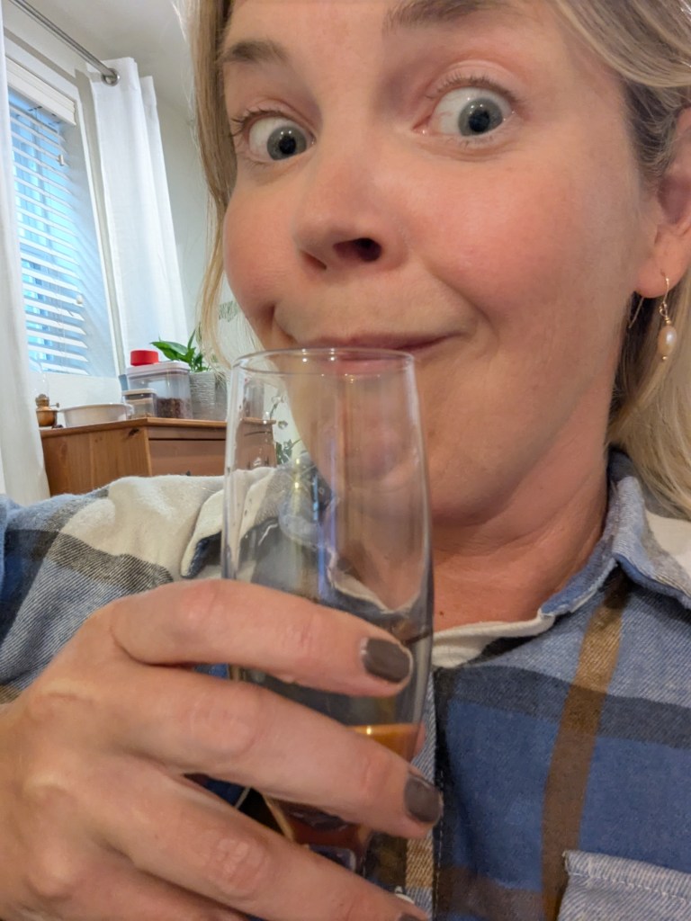 A close-up selfie of a woman holding a glass, looking playfully surprised with wide eyes and a slight smile. She is wearing a plaid shirt and has light-colored hair.