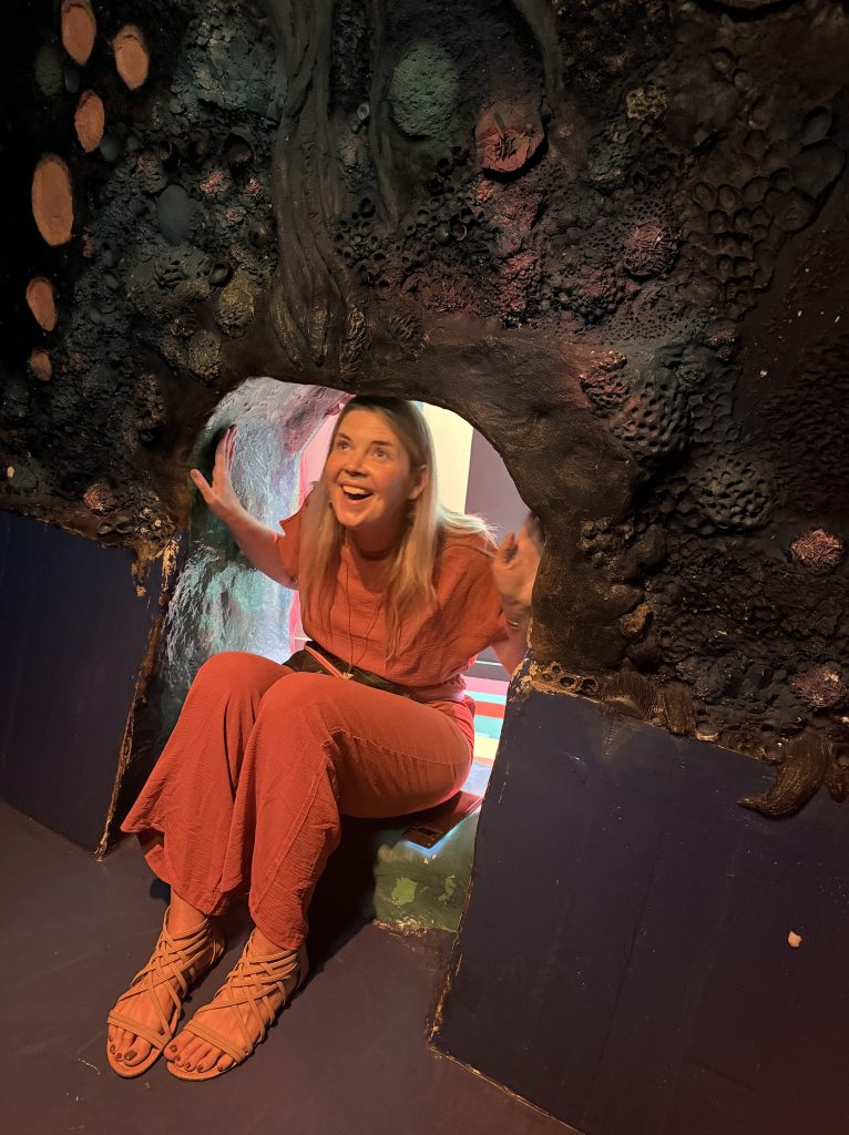 A woman in a red outfit sitting in a colorful cave-like structure, smiling and posing with her hands raised.