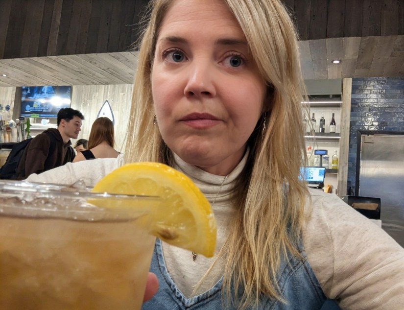 A woman holding a drink with a lemon slice, looking at the camera with a serious expression in a bar setting.