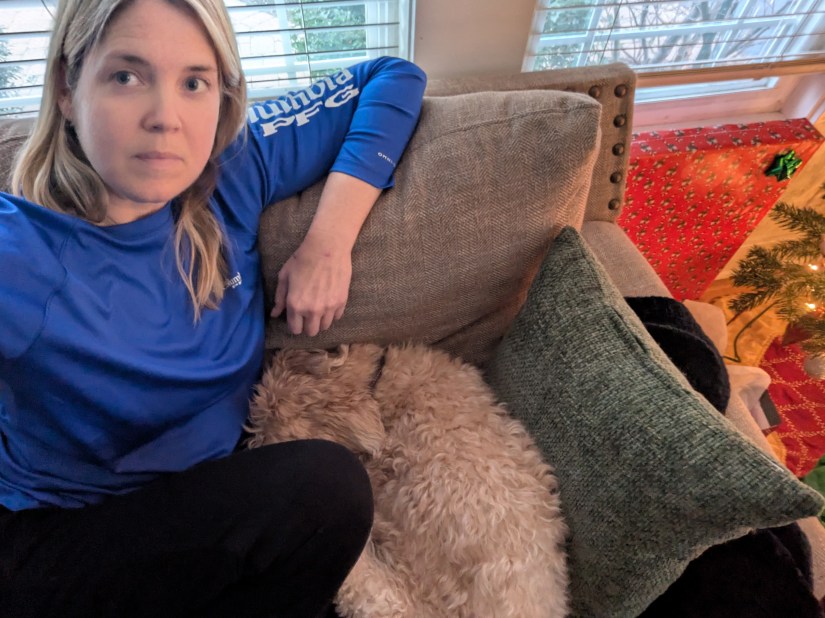 A woman sitting on a couch with a blue shirt, looking at the camera, while a fluffy dog is curled up next to her. In the background, there are festive decorations and a Christmas tree.