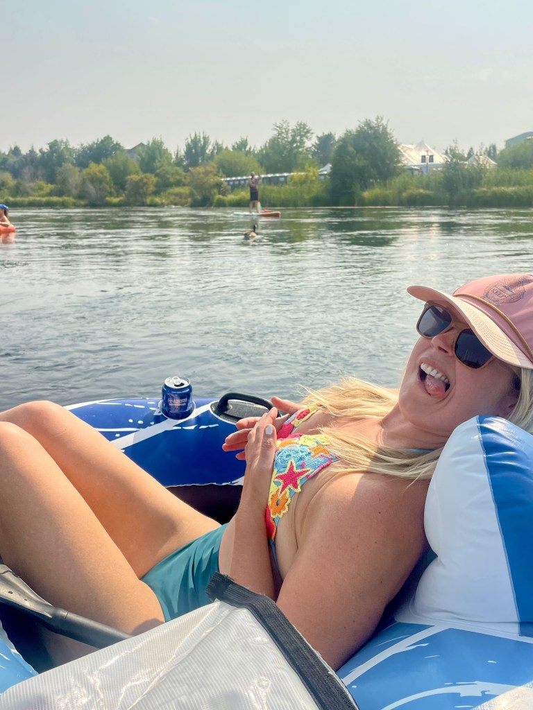 A woman relaxing in a blue inflatable raft on a river, smiling and enjoying the sun, with trees and other people in the background.
