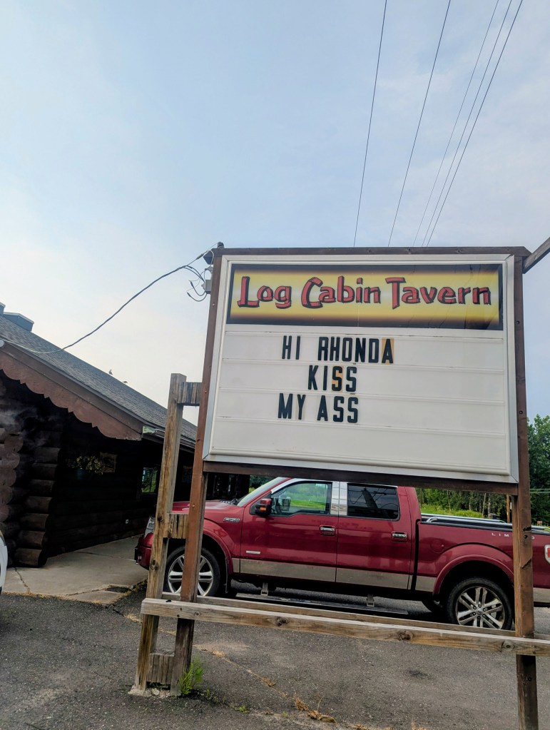 Sign outside Log Cabin Tavern with text: 'HI RHONDA KISS MY ASS'.