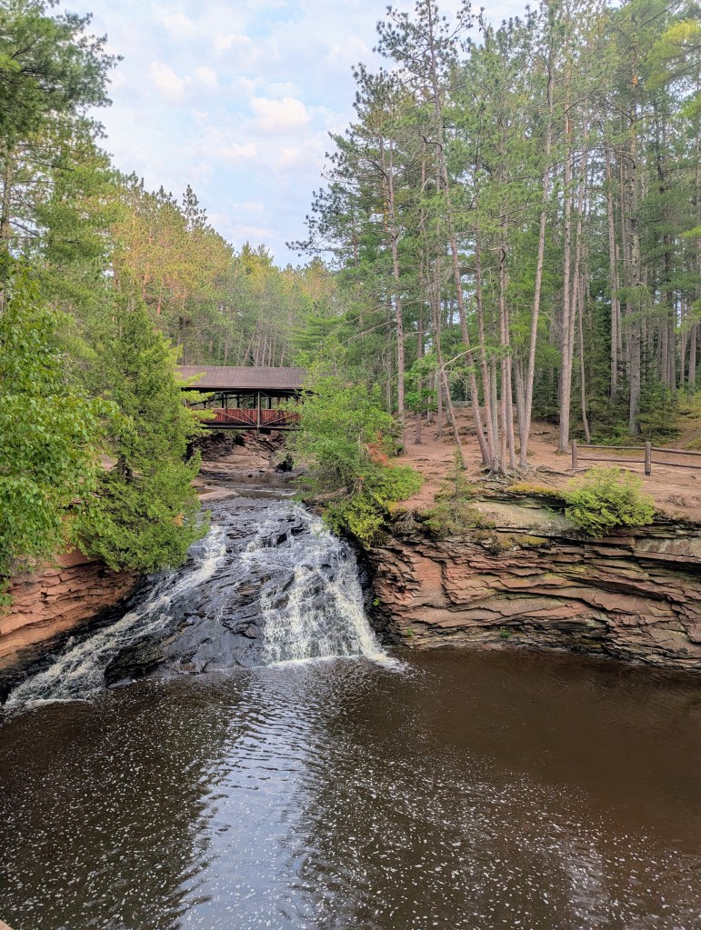 A serene landscape featuring a waterfall cascading into a tranquil river, surrounded by lush green trees and a wooden bridge in the background.