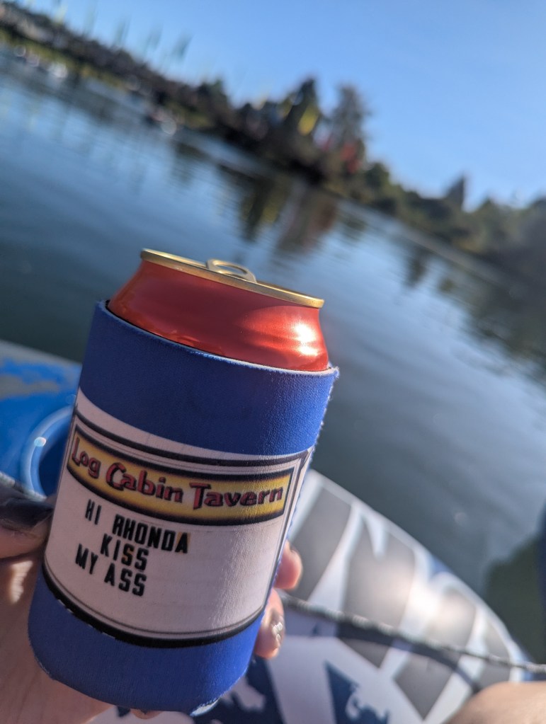 A hand holding a soda can in a blue koozie with the text 'Log Cabin Tavern Hi Rhonda Kiss My Ass', against a serene lake background with trees and blue sky.